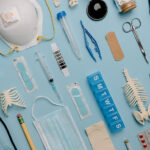 Overhead photo of medical tools arranged neatly on a sky blue background. The tools include a face mask, bandaid, pill box, tweezers, and other assorted medical implements.