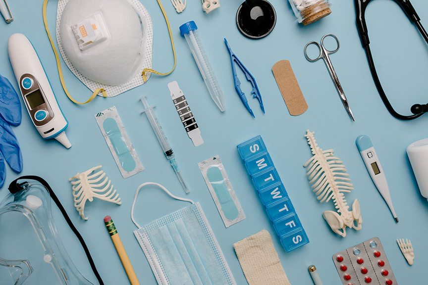 Overhead photo of medical tools arranged neatly on a sky blue background. The tools include a face mask, bandaid, pill box, tweezers, and other assorted medical implements.