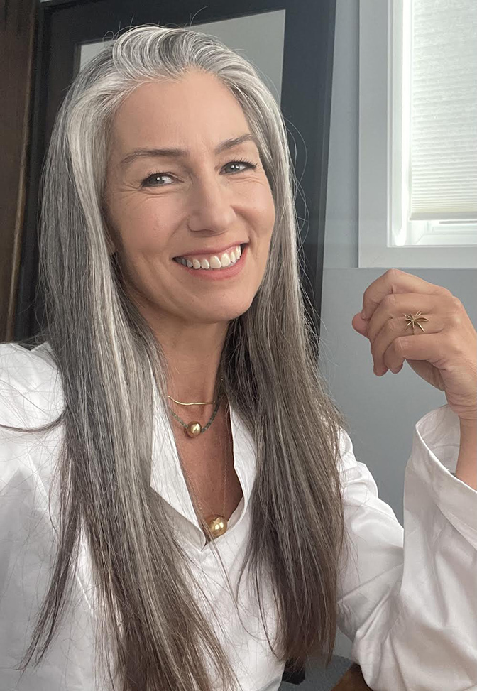 Photo of Mia Beckman, jewelry designer, posing with her long, straight grey hair and beautiful gold necklaces and ring