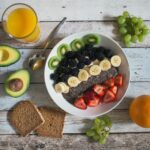 A white plate with bananas, blackberries, kiwi and strawberries on a wooden plank table with a glass of juice, two pieces of toast and a split avocado