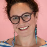 Emily McDowell, a woman wearing glasses, smiles in front of a pink background.