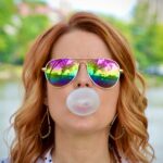 A woman wearing reflective sunglasses and blowing a bubble with bubblegum