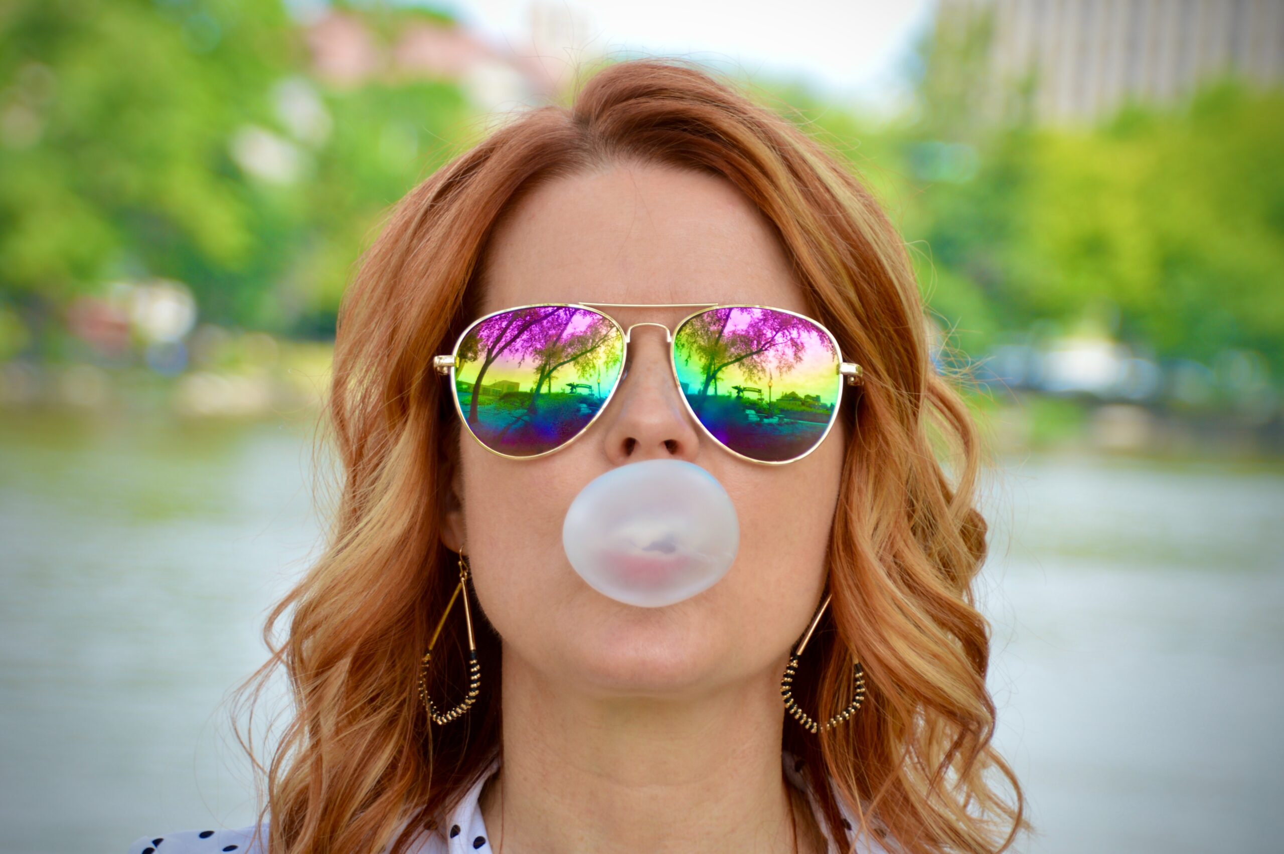 A woman wearing reflective sunglasses and blowing a bubble with bubblegum
