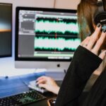 A woman's hand with painted nails holding headphones to her ear, as she records on a computer audio setup