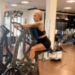 A strong woman is sitting on a stationary bike in a silver gym.