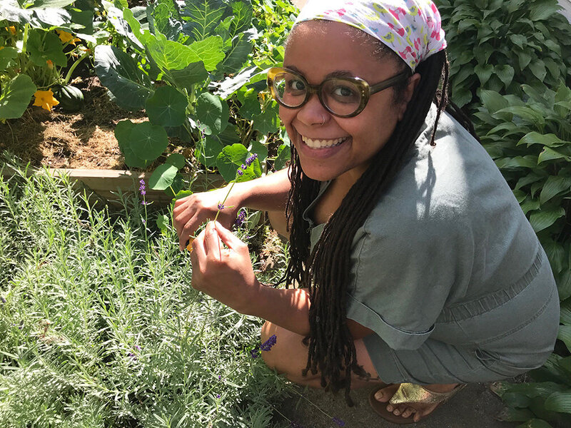 Tequia Burt in glasses kneeling in a garden as she is harvesting lavender.