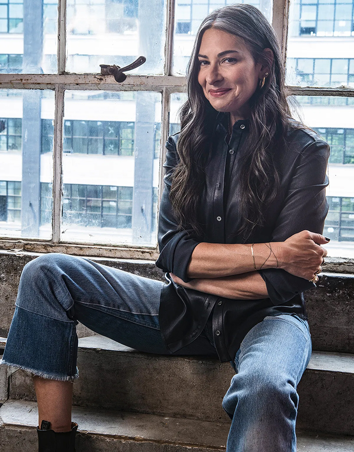 A woman sitting on a window sill wearing jeans and a leather jacket.