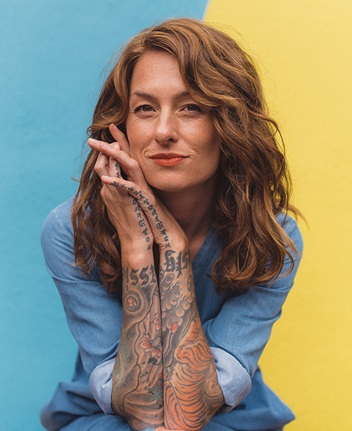 A woman with tattoos posing in front of a colorful background.