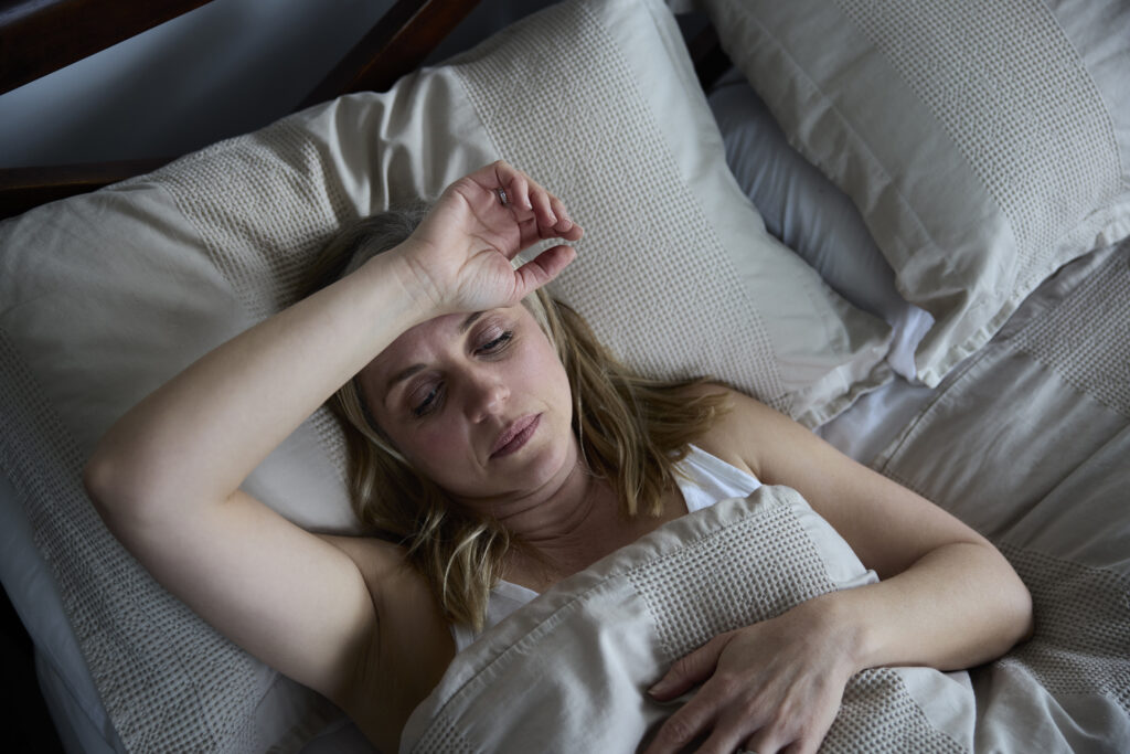 Woman with blond hair in bed looking tired, arm raised over forehead.