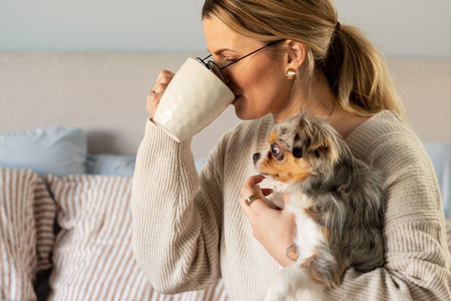 woman drinking out of cup and holding her dog on her bed