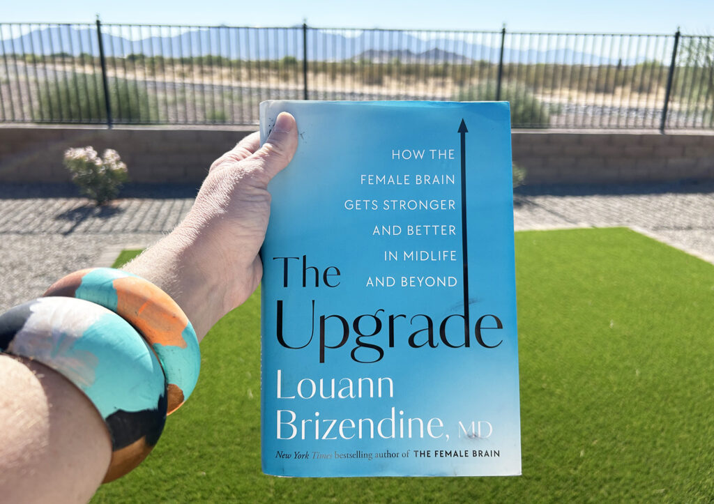 A woman holding the book called "The Upgrade: How the Female Brain Gets Stronger and Better in Midlife and Beyond" by Dr. Louann Brizendine, MD.